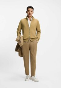 Beżowy sweter na zamek błyskawiczny z wysokim kołnierzem, w zestawieniu z jasnobrązowymi spodniami i białymi trampkami. Do tego dopasowana beżowa kurtka.