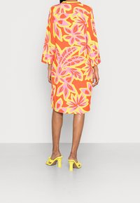 Buntes Kleid mit floraler Musterung in Pink-, Gelb- und Orangetönen, mit dreiviertellangen Ärmeln und lockerem Schnitt, kombiniert mit gelben High Heels.