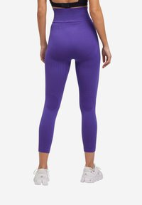 Leggings viola con un motivo testurizzato, vita alta e toppe sulle ginocchia; abbinati a scarpe sportive bianche.