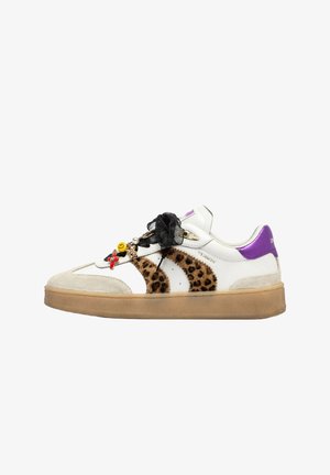 Sneaker bianco con strisce leopardate, linguetta viola, suola beige, lacci neri a nastro e dettagli decorativi tra cui una faccina sorridente e una croce.