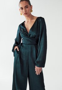 Grüner, strukturierter Jumpsuit mit tiefem V-Ausschnitt und langen, weiten Ärmeln. Verfügt über ein Wickeldesign und taillierte Taille, kombiniert mit großen goldenen Ohrringen.