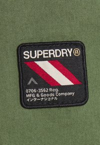 Sort lap med hvid tekst "SUPERDRY®," med røde og hvide skrå striber og en pil på en tekstureret grøn stofbaggrund.