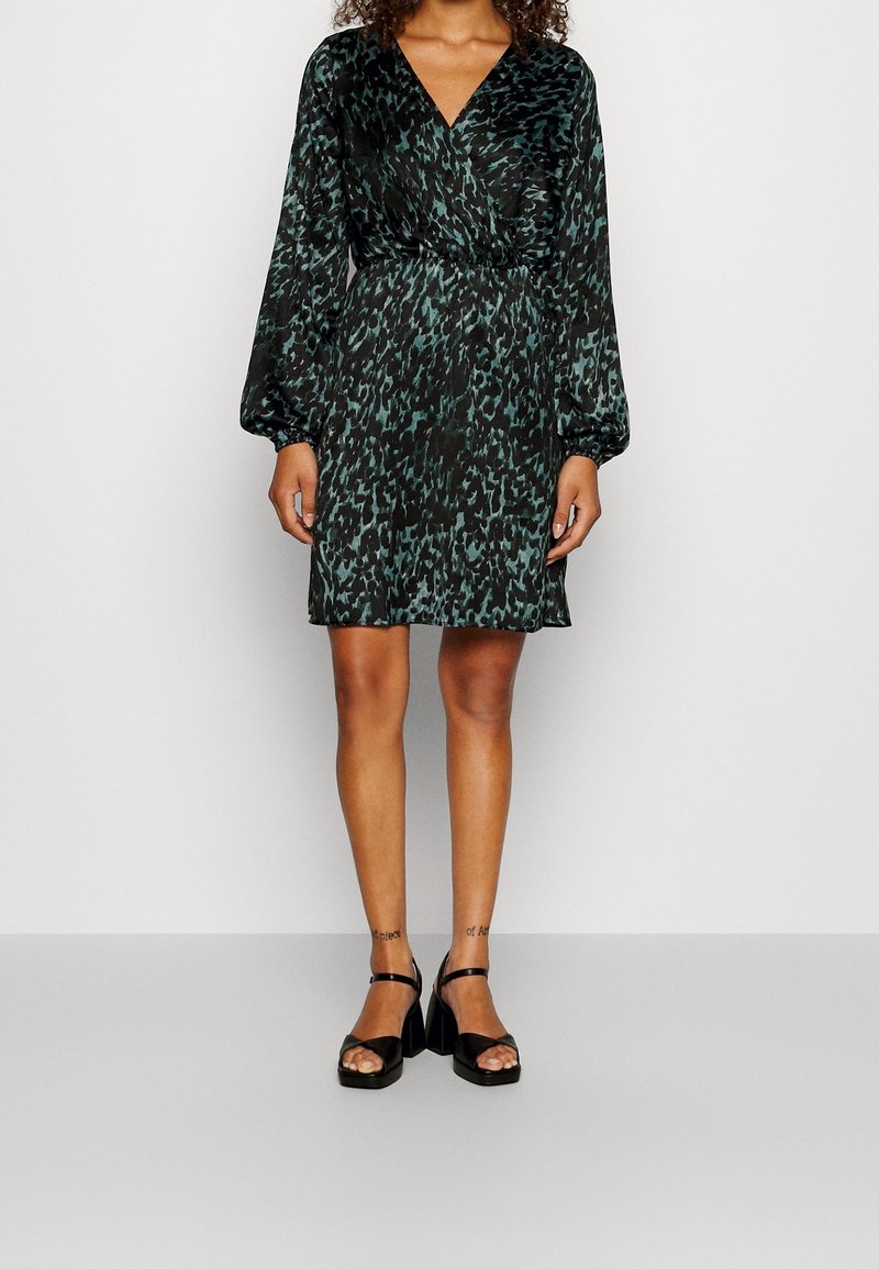 Femme portant une robe portefeuille à imprimé animalier vert foncé et noir, manches longues, et des sandales noires à talons hauts, debout devant un fond uni.