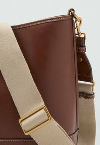 Brun lædertaske med en beige rem. Har et guld hardware-accents og kontraststing langs kanterne, hvilket fremhæver en struktureret design.