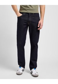 Vaqueros de mezclilla de un azul oscuro con un corte regular, cinco bolsillos y detalles de costura en contraste. Combinados con zapatillas blancas con acentos negros.
