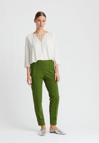 Weiße Bluse mit V-Ausschnitt und Bindegürtel, kombiniert mit grünen, taillierten Hosen mit Seitentaschen und silbernen flachen Schuhen mit strukturierter Oberfläche.