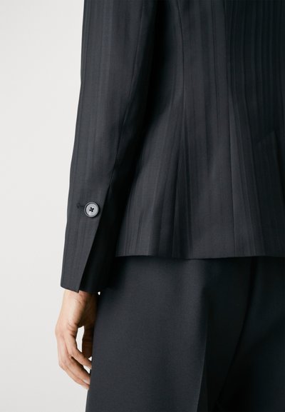 Crni blazer s prugama koji se odlikuje krojem prilagođenim tijelu, strukturiranim ramenima i detaljom jedne dugmadi na manžetama. Glatka tkanina s mat završetkom.