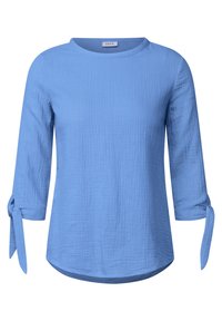 Chemise bleu clair légère avec manches aux 3/4, dotée de liens noués aux poignets et d'un ourlet légèrement arrondi. Tissu texturé.