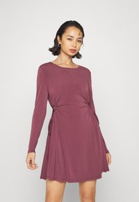 Vestido de manga longa, na cor borgonha, feito de um tecido suave, com franzidos laterais e uma silhueta a-line relaxada. Decote redondo e sem padrões.