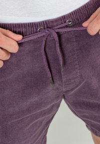 Shorts de pana morados con cintura ajustable mediante cordón, que presentan ojales de metal y una textura suave. Primer plano de la cinturilla y el detalle del cordón.