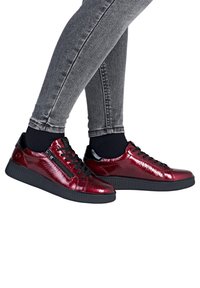 Jambes en jean skinny gris portant des baskets brillantes rouge foncé avec semelles noires, lacets, fermeture éclair latérale et chaussettes courtes noires sur fond blanc.
