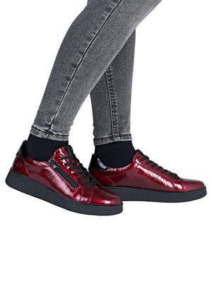 Ben i grå skinny jeans iført skinnende mørkerøde sneakers med sorte såler, snørebånd, sidelås og sorte ankelsokker på hvid baggrund.