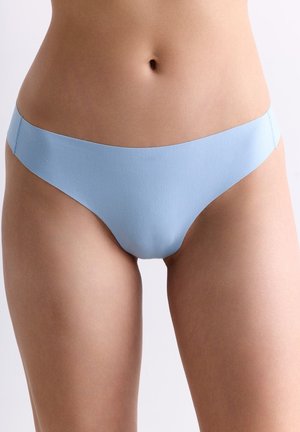 Frau trägt nahtlose hellblaue Unterwäsche, zeigt Bauchmitte und Oberschenkel vor einfachem Hintergrund.