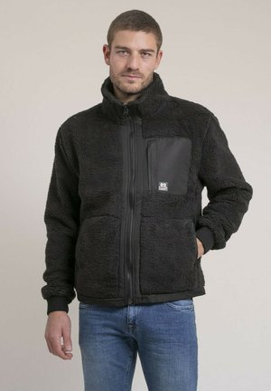 Homme portant une veste polaire noire à fermeture éclair frontale et poche poitrine, associée à un jean bleu, debout devant un fond uni.