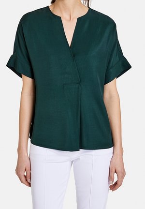 Femme portant un chemisier vert foncé à manches courtes et col en V avec un pantalon blanc, debout devant un fond clair uni.