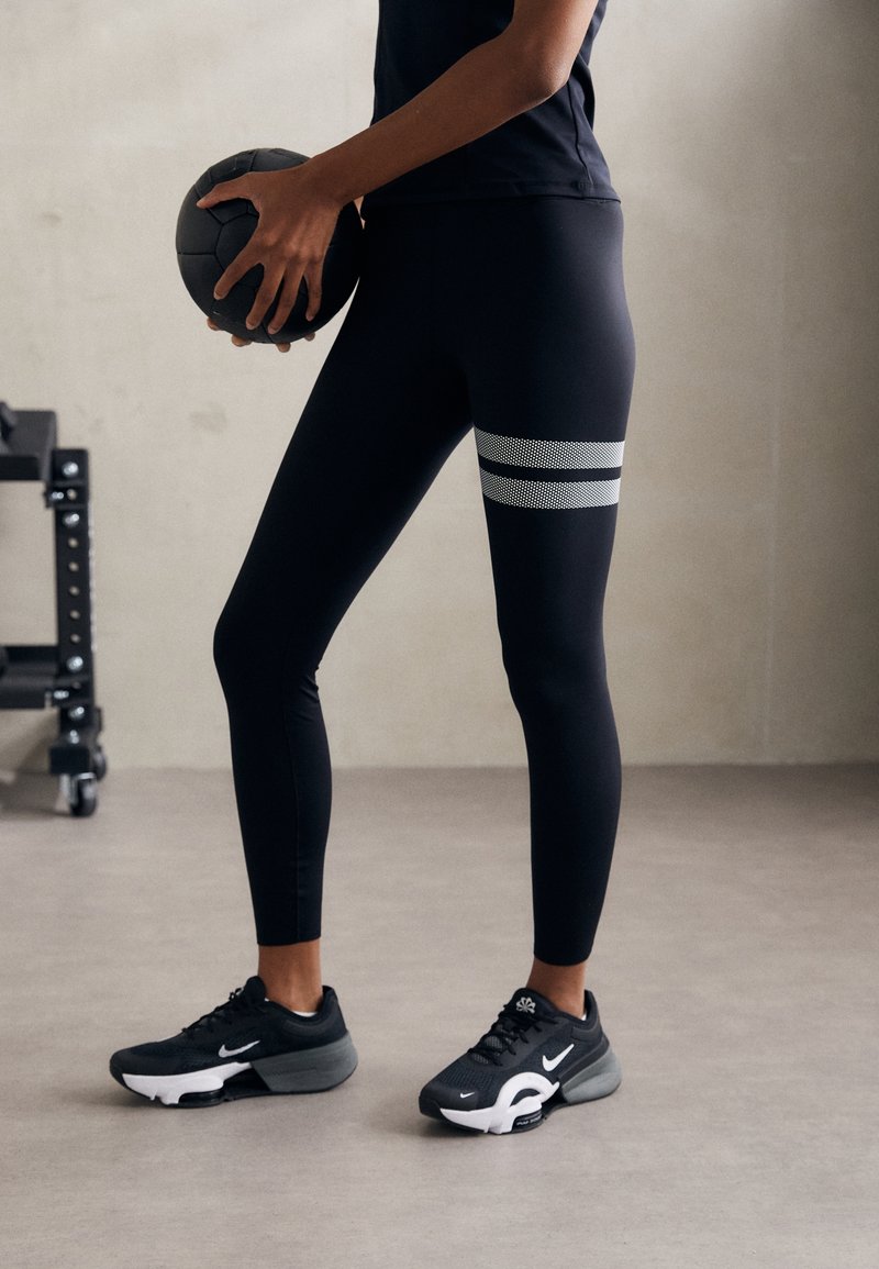 Henkilö, joka käyttää mustia leggings-housuja, joissa on kaksi valkoista raitaa, pitää mustaa kuntopalloa kädessään seisten harmaalla lattialla lähellä kuntosalivälineitä.