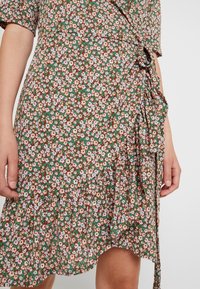 Kwiatowa sukienka w odcieniu brązowym z krótkimi rękawami, z falbanistym dołem, małymi różowymi i niebieskimi kwiatkami na zielonym tle oraz związaną z boku.