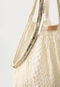 Kremowa torba na ramię z plecionym wzorem siatki, z tkaninowymi paskami ozdobionymi cekinami oraz brązową skórzaną metką z napisem "VANESSA BRUNO".