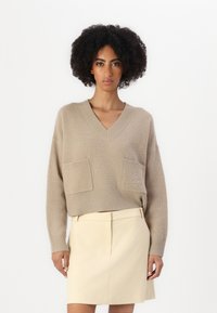 Maglione beige lavorato a maglia con scollatura a V, due tasche frontali e una texture sottile. Indossato con una gonna su misura di colore chiaro.