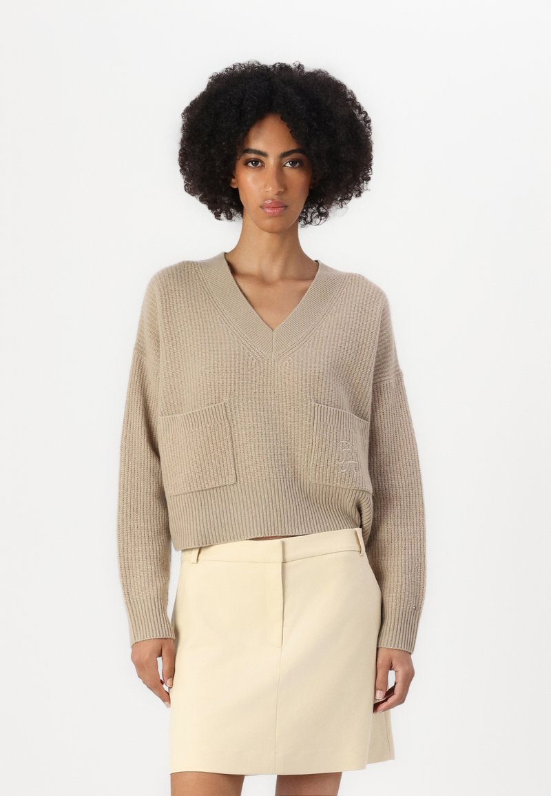 Maglione beige lavorato a maglia con scollatura a V, due tasche frontali e una texture sottile. Indossato con una gonna su misura di colore chiaro.