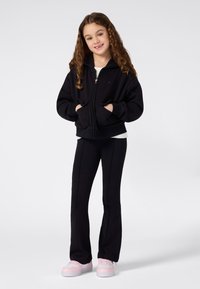 Niña joven con cabello rizado largo, sonriendo y vestida con una sudadera negra con cremallera, pantalones negros y zapatillas rosas, sobre un fondo blanco.