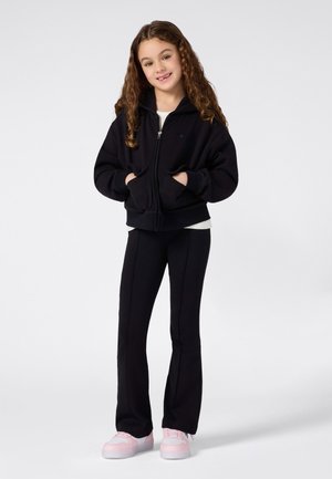 Ragazza giovane con lunghi capelli ricci, sorridente, indossa una felpa nera con zip, pantaloni neri e sneaker rosa su uno sfondo bianco.