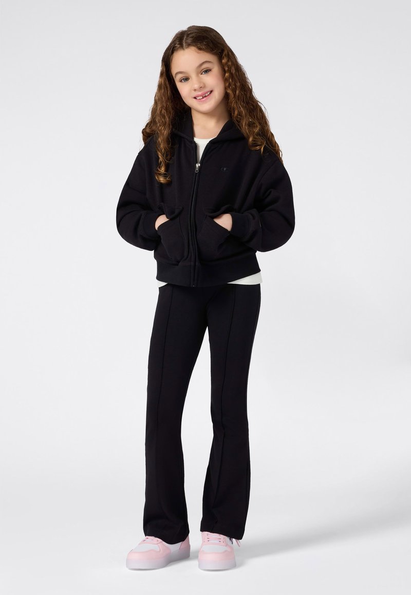 Niña joven con cabello rizado largo, sonriendo y vestida con una sudadera negra con cremallera, pantalones negros y zapatillas rosas, sobre un fondo blanco.