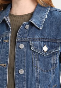 Veste en denim bleu avec une coupe classique. Présente des boutons en métal, deux poches poitrine et des accents usés sur les bords.