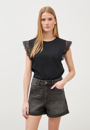 Frau mit glattem blondem Haar trägt ein schwarzes ärmelloses Oberteil mit Spitzenärmeln und hoch taillierte, aufgerollte schwarze Jeansshorts, steht vor einem einfarbigen Hintergrund.