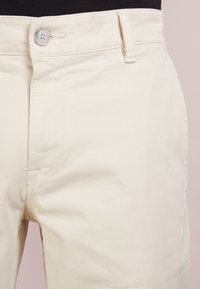 Pantalon en coton beige avec une texture lisse, doté d'une fermeture à boutons sur le devant, de passants pour ceinture, et de détails de poches latérales.