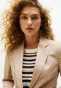 Blazer beige con Rever strutturati, indossato sopra un top in maglia a fantasia con strisce nere, bianche e blu navy. Capelli ricci che incorniciano il viso.