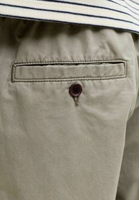 Pantalon kaki avec une poche arrière fermée par un bouton marron. Confectionné en tissu lisse présentant une texture subtile et des détails de couture.