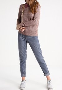 Hellrosa gestrickter Pullover mit strukturierter Oberfläche, kombiniert mit hochgekrempelten blauen Jeans und weißen Turnschuhen, ergänzt durch eine beige Umhängetasche.