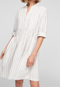 Robe légère à rayures avec une patte de boutonnage, un col, deux poches poitrine et des manches retroussées dans des tons doux de beige et de blanc.