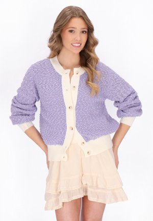Ung kvinde med bølget hår iført en lavendel tekstureret cardigan med cremefarvet kant og et lyst cremefarvet lagdelt skørt, smilende med hænderne på hofterne.