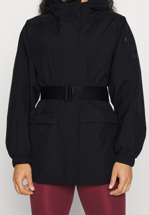 Person trägt eine schwarze Jacke mit Gürtel, großen Fronttaschen und elastischen Bündchen, kombiniert mit roten Leggings.