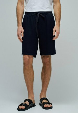 Hombre con pantalones cortos negros con cordón y sandalias negras con hebilla, de pie frente a un fondo gris.