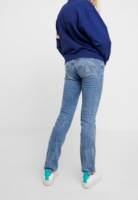 Vaqueros azules de corte ajustado y textura desgastada, combinados con una sudadera azul marino que presenta una franja multicolor en la manga. Zapatillas blancas con acentos en azul turquesa.