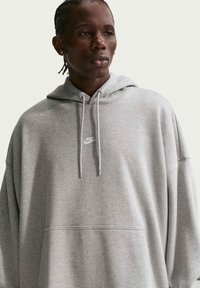 Homme aux cheveux tressés portant un sweat à capuche Nike gris avec cordons, regardant légèrement sur le côté sur un fond uni.