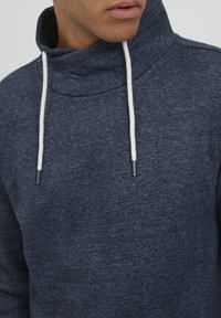 Sötétkék kapucnis pulóver, magas gallérral, fehér zsinórokkal és struktúrált anyaggal. Nagyítás a nyakrészre és az arcfelületre.