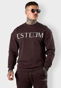 Brauner Sweatshirt mit weißem Text "ESTEEM", mit gerippten Bündchen und Saum, kombiniert mit passenden Hosen. Glatte Stoffoberfläche.