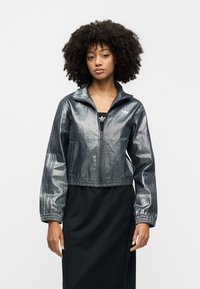 Metalno siva kraća jakna s patentnim zatvaračem sprijeda, visokim ovratnikom i elastičnim manšetama. Ima mali bijeli logo na lijevoj strani prsa.