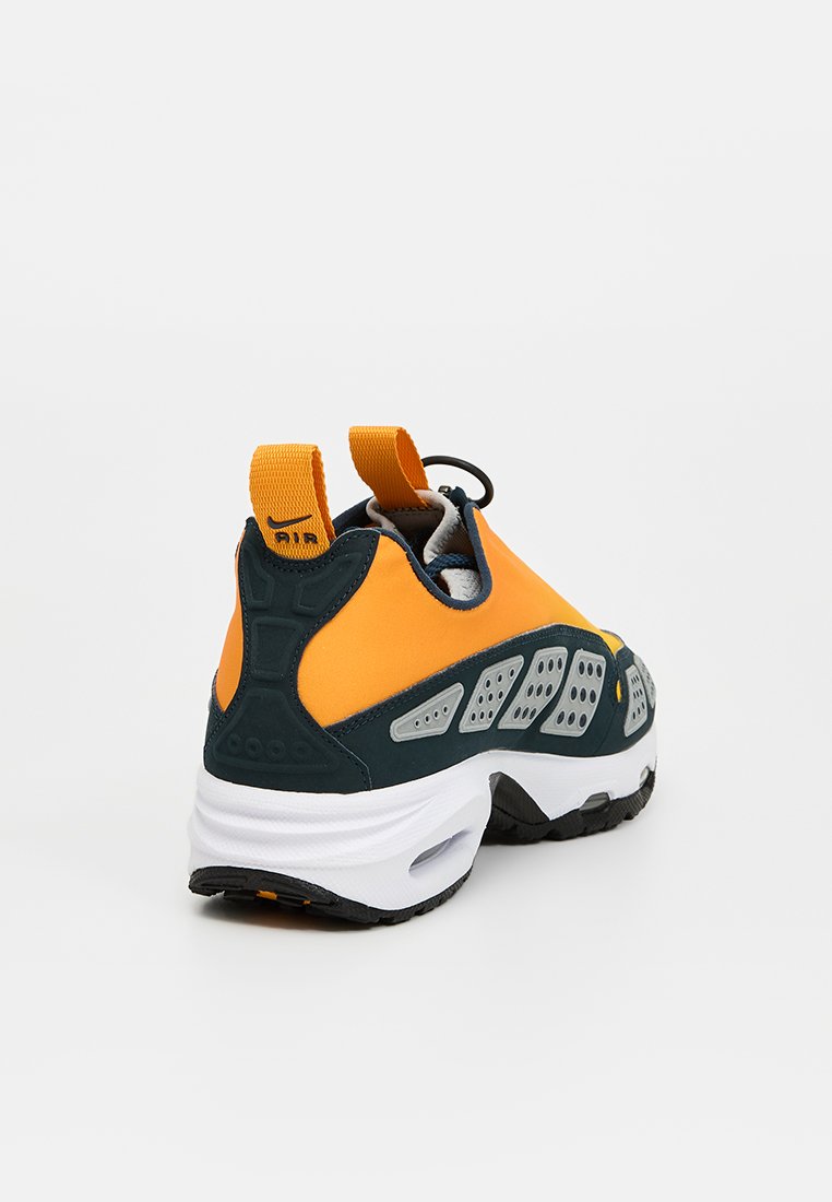 Zapatilla deportiva con parte superior naranja y acentos grises, suela negra y detalles texturizados; incluye un tirador en la parte trasera para facilitar su uso.