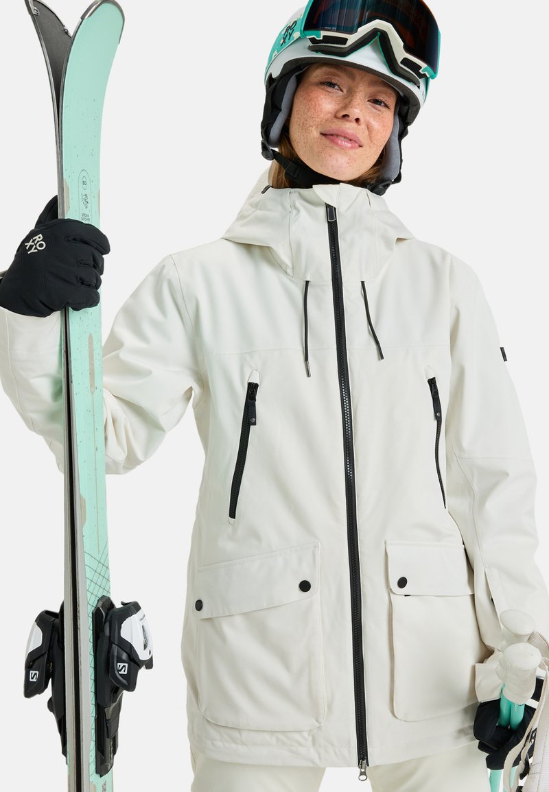 Weiße Skijacke mit schwarzen Reißverschlüssen, verstellbarer Kapuze und zwei Fronttaschen. Die Person hält Skier und trägt einen Helm sowie eine Skibrille.