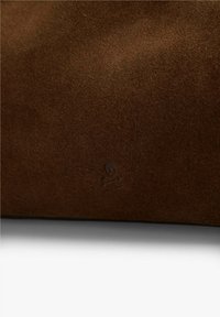 Material de ante marrón con una textura suave. Presenta un sutil grabado de calavera y huesos cruzados. El diseño es minimalista y sin costuras.