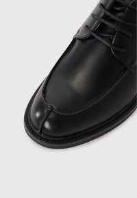 Chaussure en cuir noir avec un bout arrondi, texture lisse, détails cousus et design à lacets. La semelle présente une légère élévation.