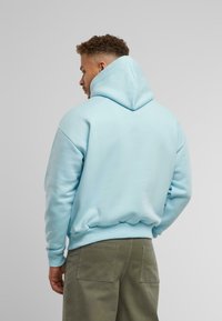 Lichtblauwe hoodie met een grote capuchon, geribde mouwen en taille. De stof lijkt zacht en glad. Draagt met olijfgroene broeken.