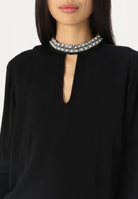 Blouse noire avec un décolleté en clé et un col orné de perles et d'accents argentés, conçue avec des manches drapées pour une coupe décontractée.