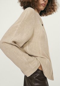 Beige linned sweater med løse ærmer, sideslidser og en enkelt mørk knaplukning. Båret over mørke læderbukser.
