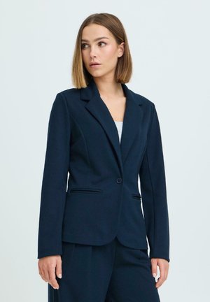 Õlgadeni juustega naine, kes kannab keha järgi sobivat tumesinist bleiserit ja sobivaid pükse, vaatab valgusküllase tausta taustal küljele.